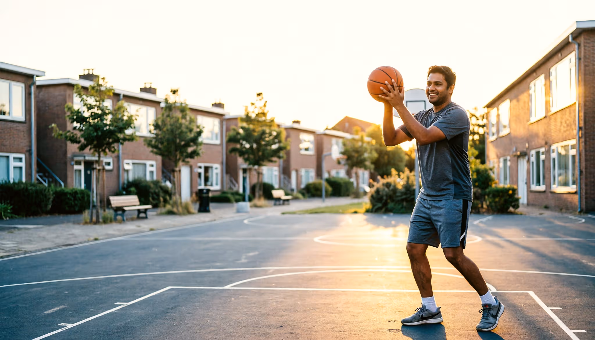 A young man shoots hoops on an outdoor basketball court in a Dutch neighbourhood at golden hour