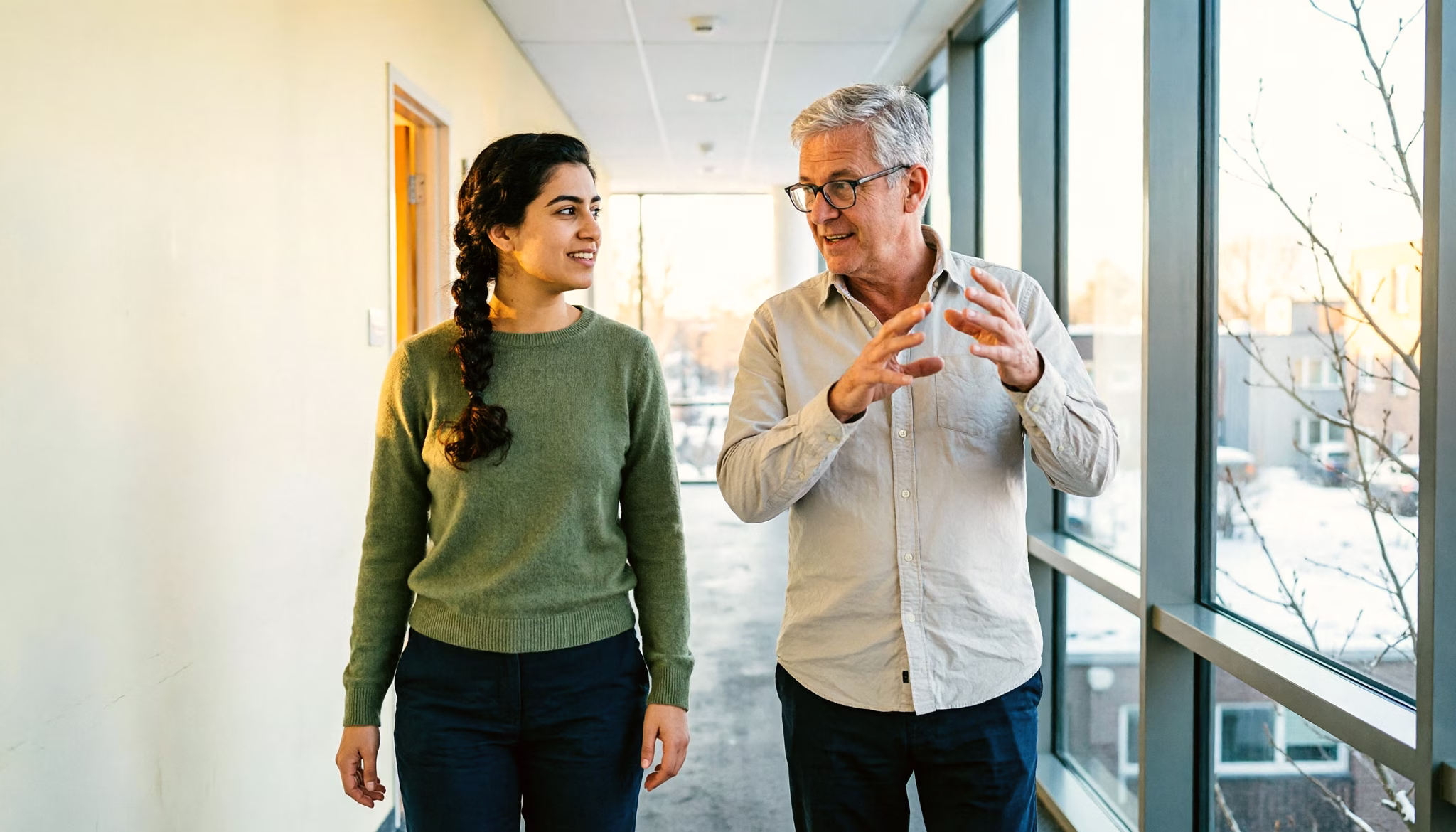 Een ervaren collega legt een klantsituatie uit aan een jongere collega terwijl ze door een lichte kantoorgang lopen