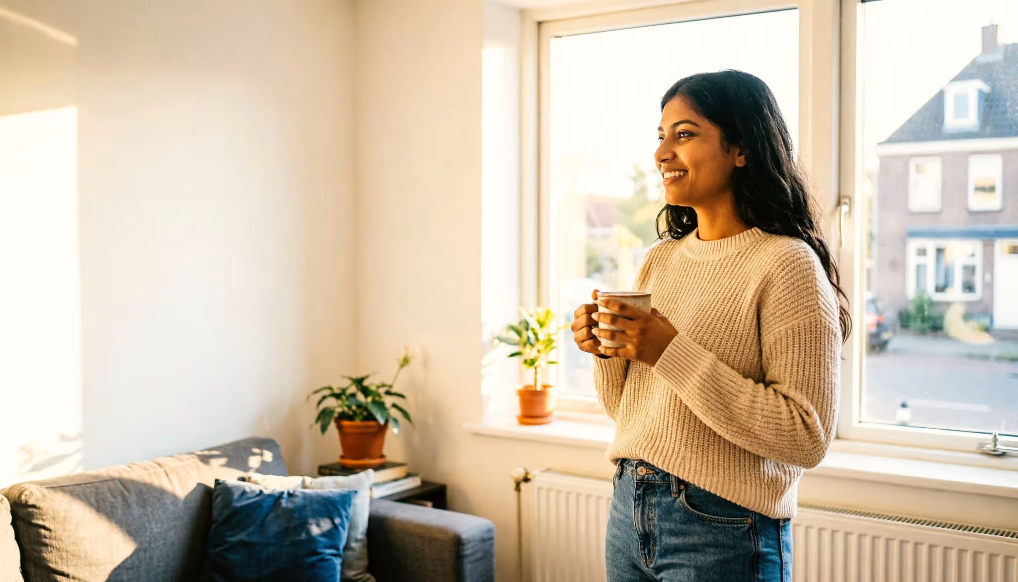 Een vrouw staat met een kop thee voor het raam van haar appartement en kijkt uit over een Nederlandse straat