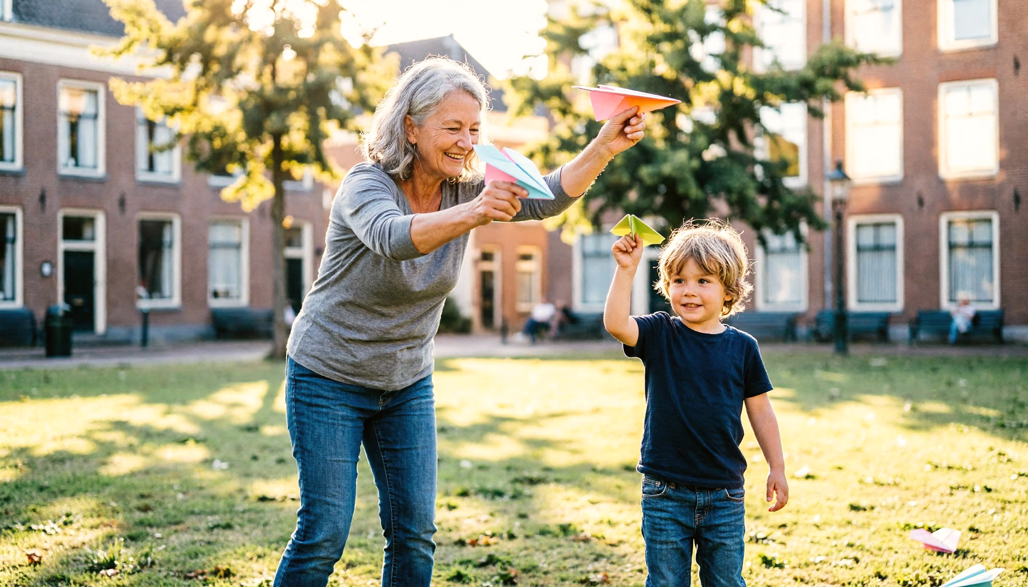 An older woman and her young grandchild launch folded paper airplanes across a sunny park lawn