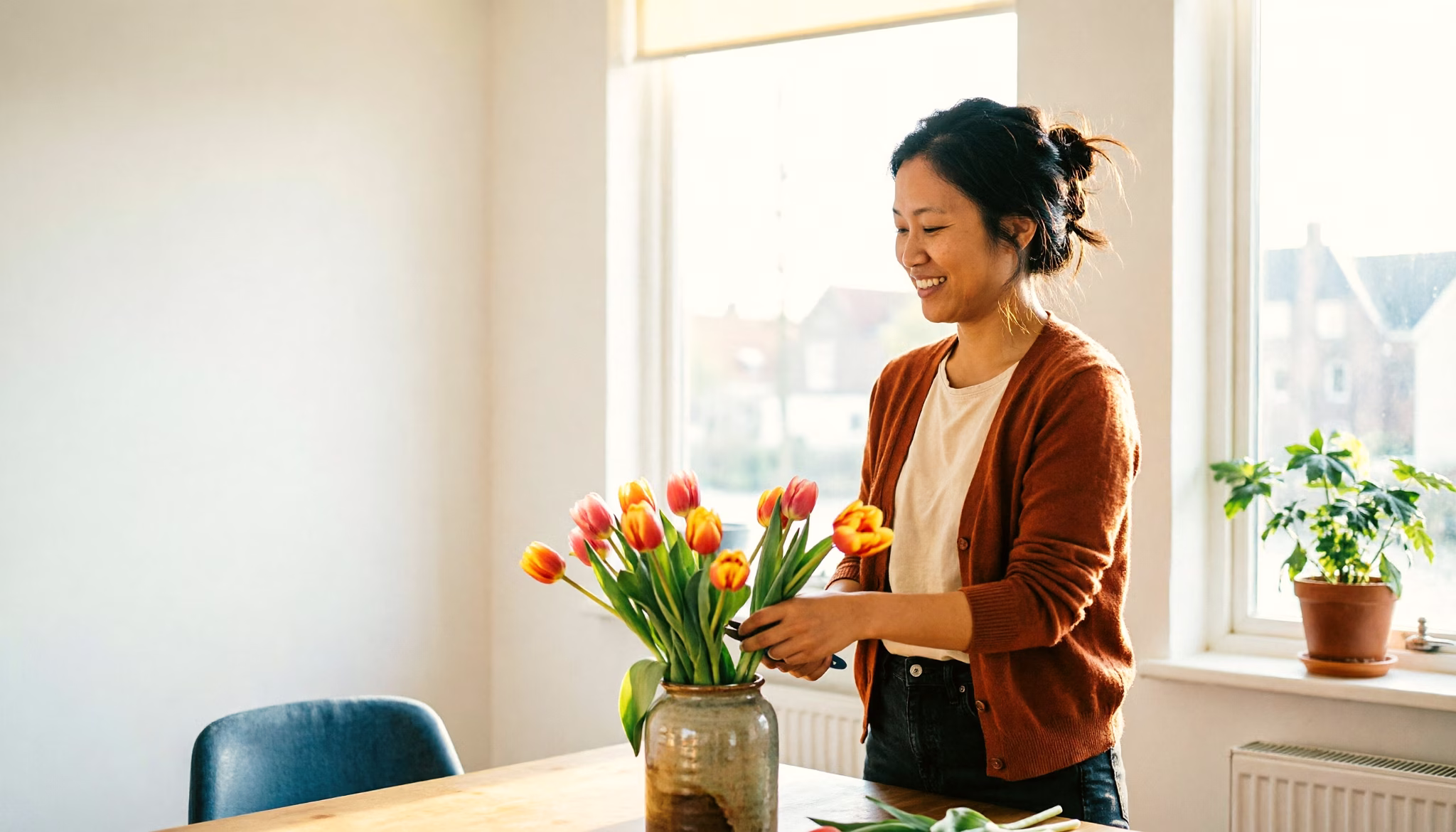 A woman in her mid-thirties arranges a bunch of tulips into a vase on the table of her sunny flat.