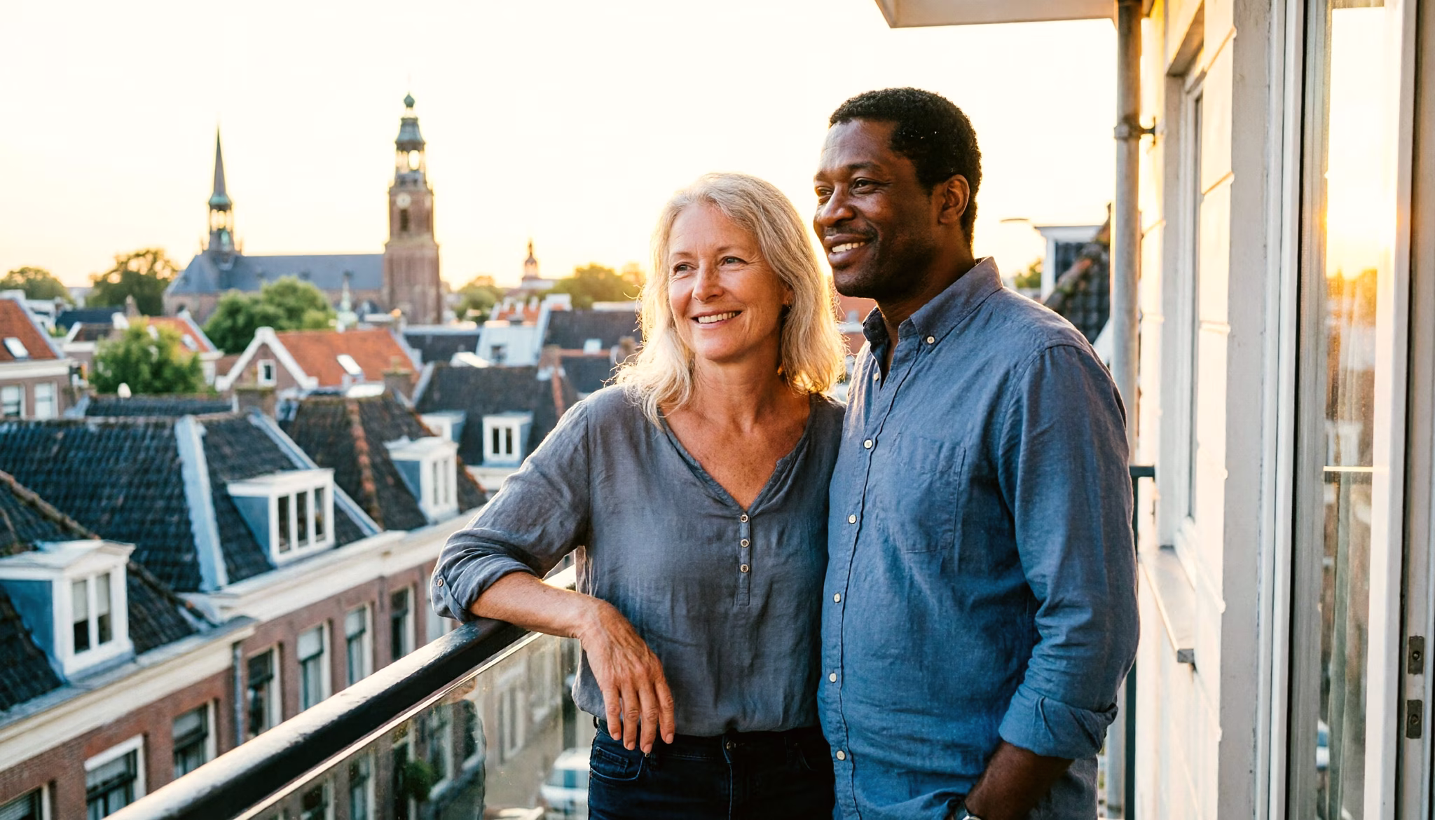 A couple in their mid-fifties stands on a balcony overlooking a Dutch cityscape, relaxed